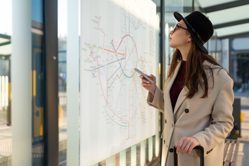 Foto de mulher em ponto de ônibus olhando a tela do celular e o mapa físico de linhas de ônibus, como é característico em uma smart city.