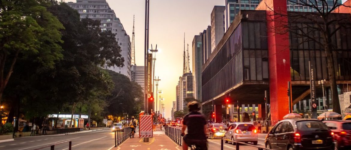 Foto de ciclista andando de bicicleta na ciclovia da Avenida Paulista, em São Paulo, próximo a outros carros em movimento.